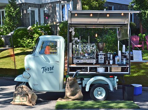 Pale blue three-wheeled vehicle with a coffee bar on its back under an awning, sacks of beans in front. Opens a larger view