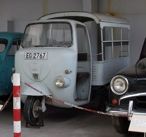 Pale grey three-wheeled minibus with open sides behind a red and white post and chain.
