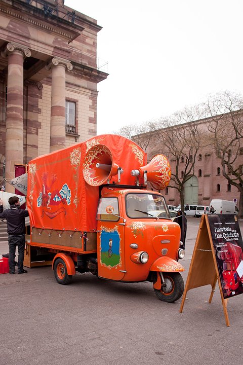 Orange three-wheeled van with large horn loudspeakers on its roof parked on a square by columns.
