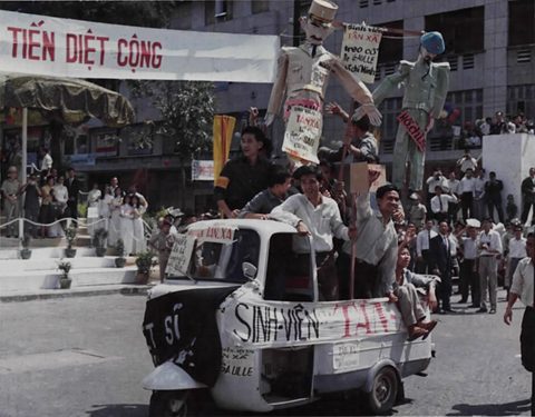 A white three-wheeled vehicle covered in slogans carries students and two effigies past a crowd.
