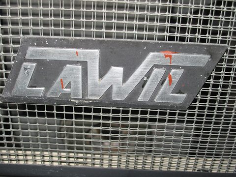 Brushed metal LAWIL letters on a black plate, set in a square wire mesh grille, with flecks of red paint. Opens a larger view