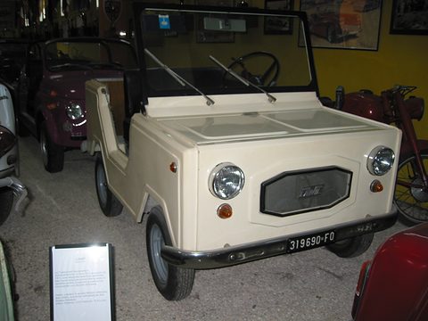 Cream open two-seater with an upright windscreen, a grille panel and round headlamps, in a crowded museum hall. Opens a larger view