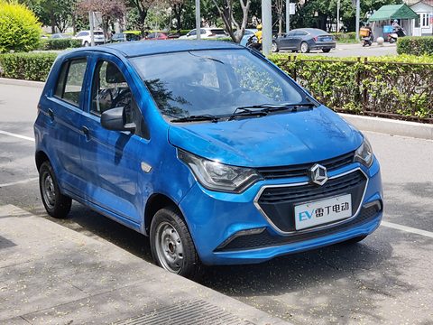 Blue small four-door hatchback with a chrome grille and EV plate parked on a pavement beside a hedge.