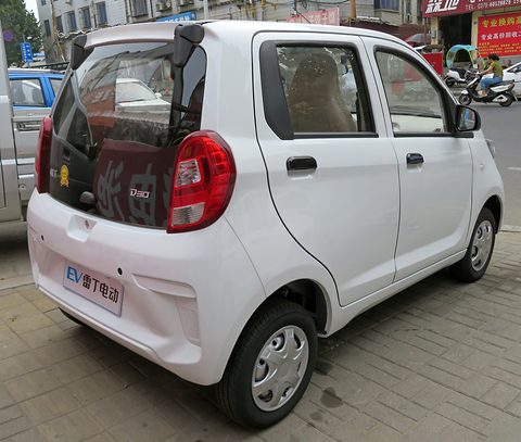 Rear of a white small four-door hatchback with an EV badge in place of a plate, on a pavement by shops. Opens a larger view