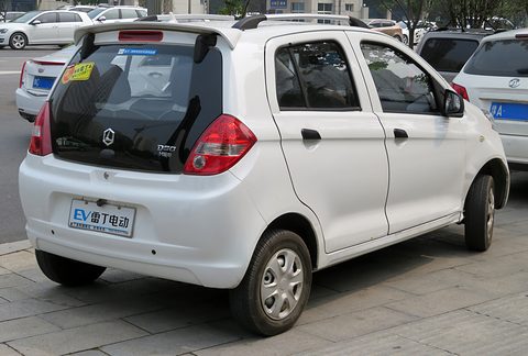 Rear of a white small four-door hatchback with roof rails and an EV plate parked on a paved pavement. Opens a larger view