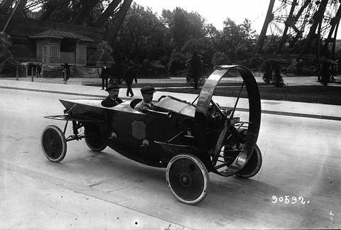A long, narrow open car with a big ringed propeller at its nose and two men in tandem, under the iron legs of the Eiffel Tower. Opens a larger view