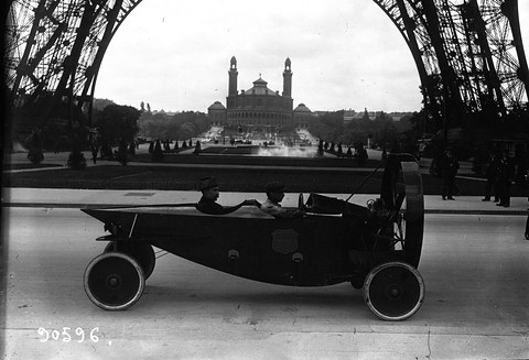 Side view of a long boat-shaped car with a ringed propeller at its front, two men aboard, framed by the arch of the Eiffel Tower. Opens a larger view