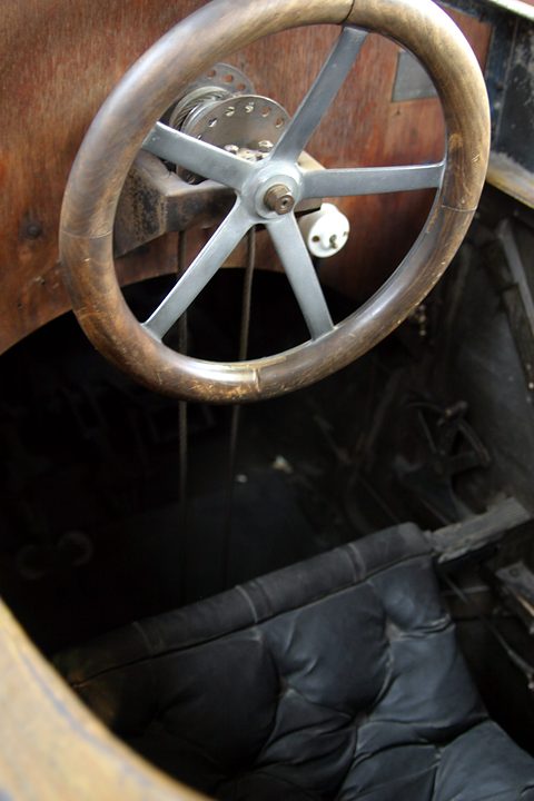 A wooden-rimmed steering wheel with metal spokes above a black buttoned seat inside a bare plywood body.