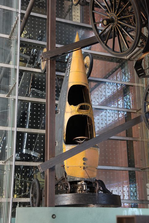 A pointed yellow plywood car body with two open cockpits, stood on end inside a tall glass and steel display tower. Opens a larger view