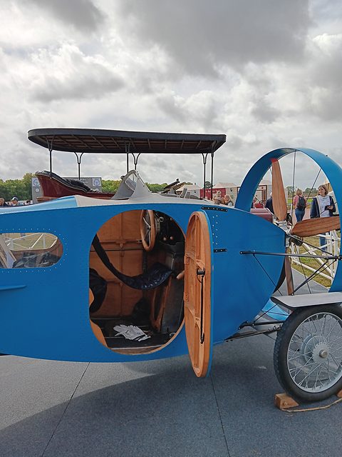 A blue riveted car body with a round-topped wooden door open on a tan cockpit, a propeller ring beyond. Opens a larger view