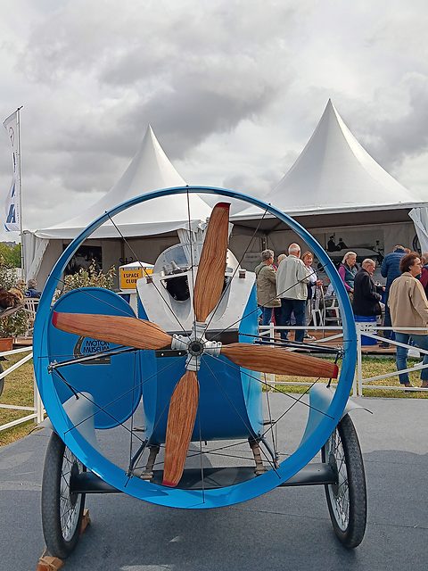 Front view of a car fronted by a large wooden propeller inside a blue ring, visitors and white marquees behind. Opens a larger view