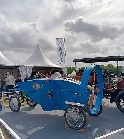 A blue fuselage-shaped car on four spoked wheels with a ringed propeller at its nose, beside a veteran car under a canopy. Opens a larger view