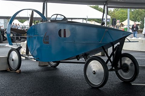 Side view of a blue riveted fuselage on four disc wheels, a ringed propeller at the front and a small round cockpit. Opens a larger view