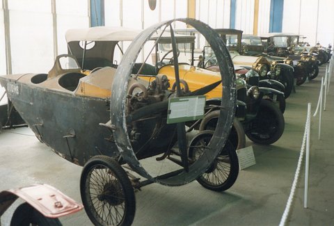An unrestored grey plywood propeller car with a large metal guard ring and wire wheels beside a row of old cars. Opens a larger view