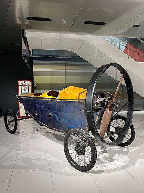 A slender blue plywood car with a yellow deck, two cockpits and a two-bladed wooden propeller ringed at its front. Opens a larger view