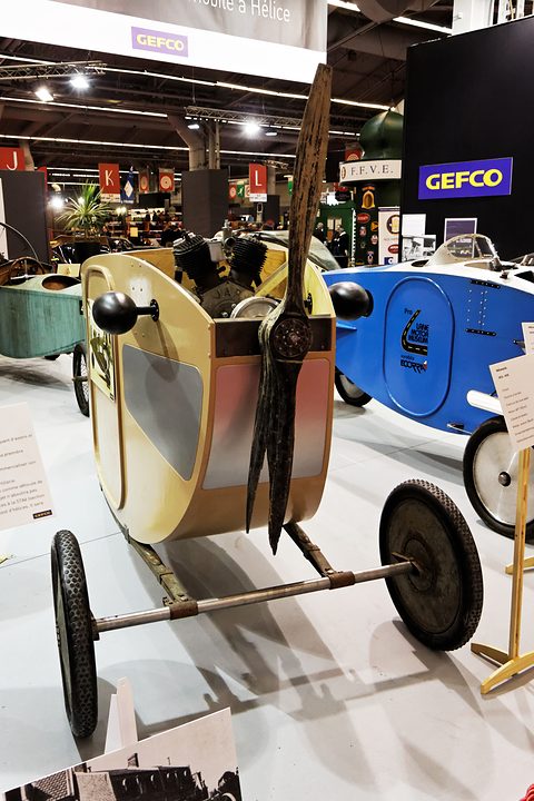 A small cream and pink cockpit body on spindly wheels with a V-twin and a long two-bladed wooden propeller in front. Opens a larger view