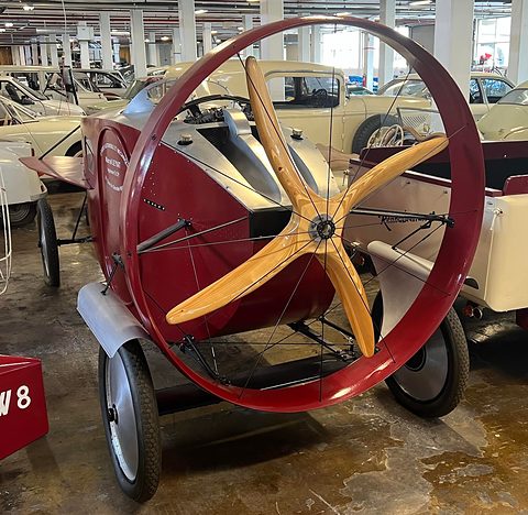 A red propeller car with a large red guard ring and a five-bladed wooden propeller, cars parked behind in a hall.