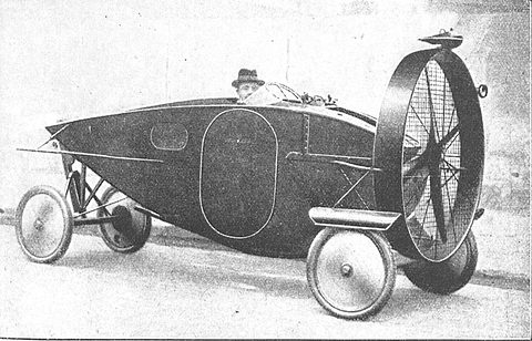 A slim plywood car body on four disc wheels, a large caged propeller at one end and a man in a hat in the cockpit.