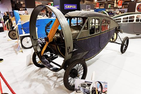 An enclosed propeller car with a navy and grey body, small windows, a wooden propeller in a guard ring and wire wheels.