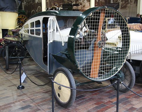 A grey and green enclosed propeller car with many small windows and a mesh-covered propeller ring, on a brick floor. Opens a larger view