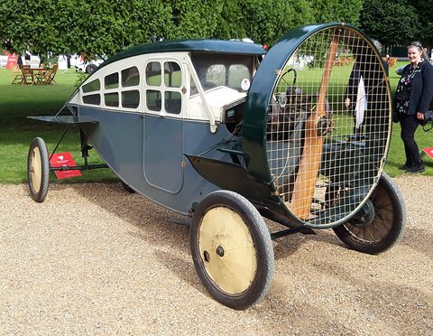 A grey-blue enclosed propeller car with small square windows and cream disc wheels on gravel, a spectator beside it. Opens a larger view