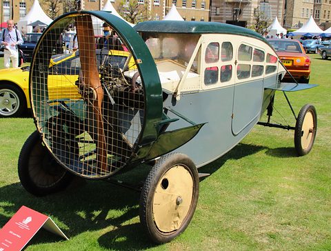 A grey-blue enclosed propeller car with a mesh-guarded wooden propeller and cream disc wheels on a lawn. Opens a larger view