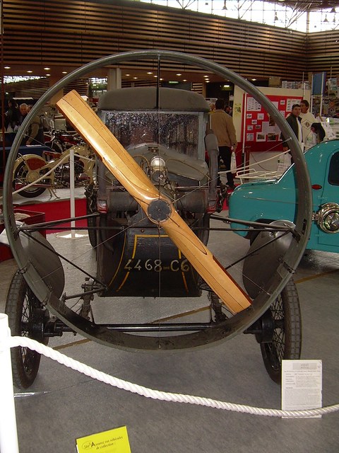 Front view of a narrow enclosed car behind a large wooden propeller set in a thin metal ring, roped off in a show hall. Opens a larger view