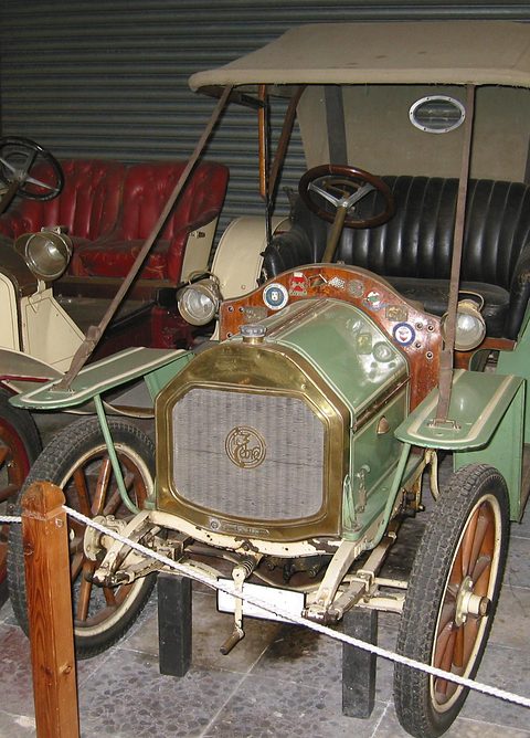 Green two-seater with a tan hood, brass radiator and badges, behind a rope barrier. Opens a larger view