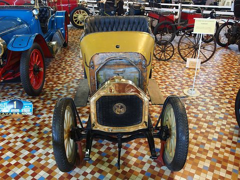 Front of a small yellow car with a brass radiator, script roundel and brass lamps on a tiled floor. Opens a larger view