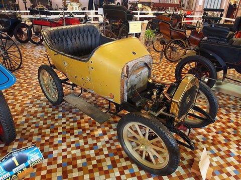 Yellow single-cylinder two-seater with black seats and wooden wheels on a chequered tile floor. Opens a larger view
