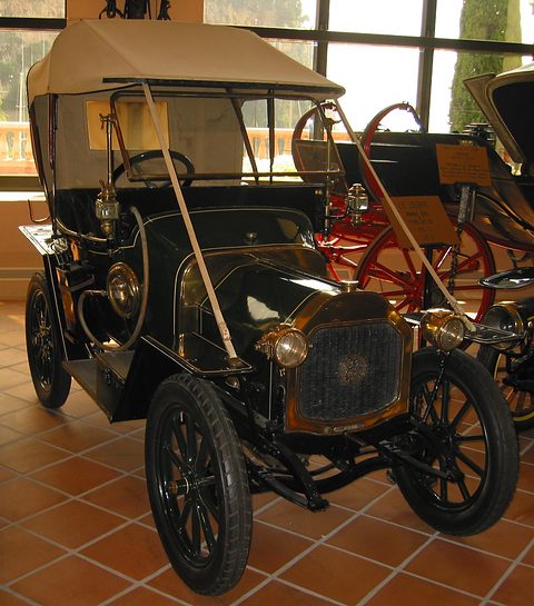Black two-seater with a cream hood, brass lamps and black wheels on a terracotta tiled floor. Opens a larger view