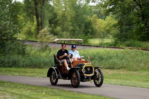 A small veteran car with a fringed canopy driving along a path between grass and trees. Opens a larger view