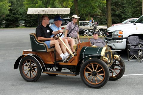 Side view of a veteran car with wooden wheels and a fringed canopy, men aboard and beside it. Opens a larger view