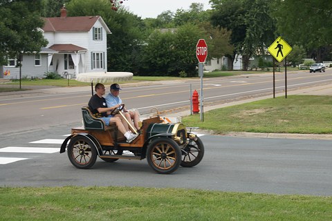 Small veteran car with a fringed canopy and two men aboard crossing a suburban road junction. Opens a larger view