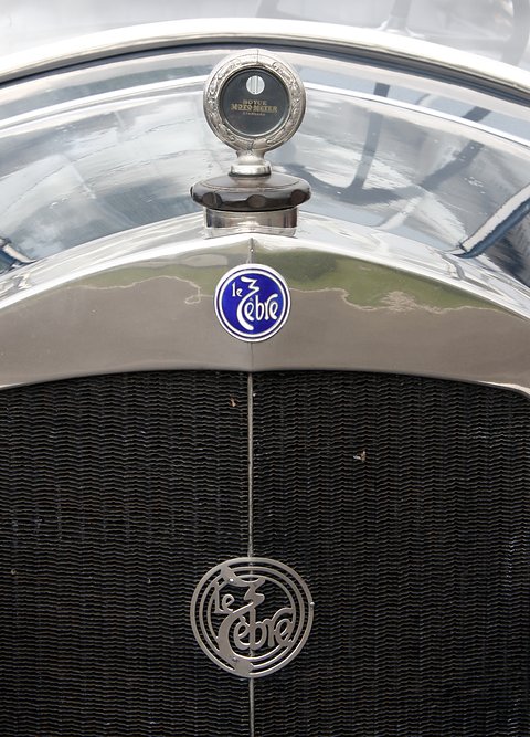 Close view of a chrome radiator top with a temperature gauge cap and a blue script badge. Opens a larger view