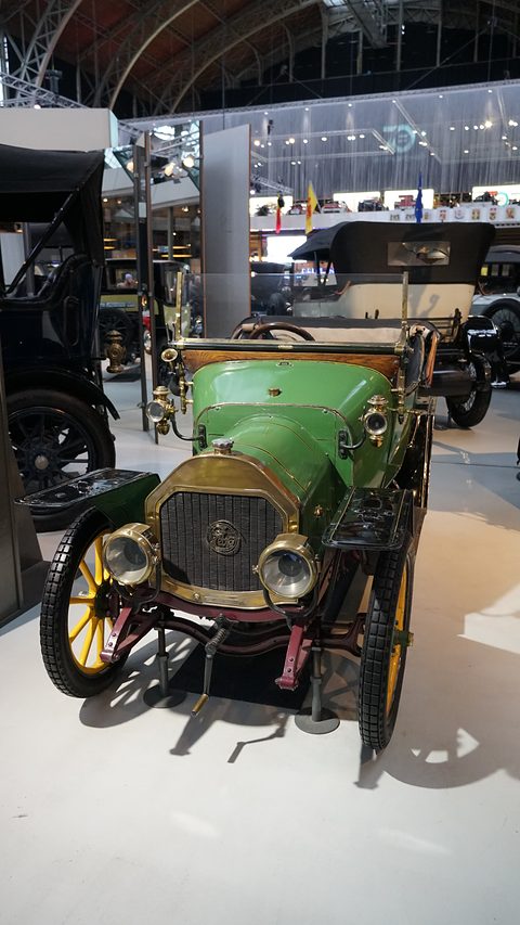 Green two-seater with brass radiator and lamps and yellow wheels in a large museum hall. Opens a larger view