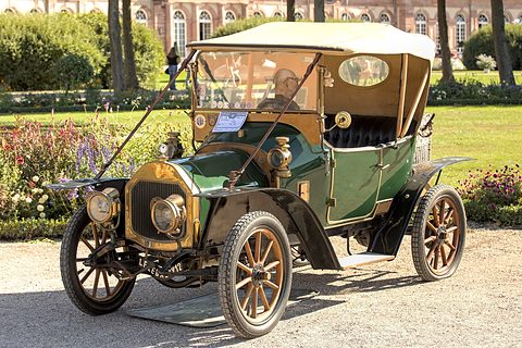 Green two-seater with a cream hood and brass radiator on gravel before palace gardens. Opens a larger view