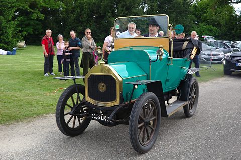 Turquoise two-seater with brass radiator and wooden wheels on a lane, people watching from the grass. Opens a larger view