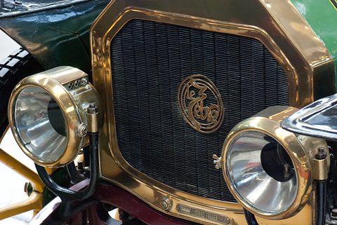 Close view of a brass radiator with a script badge between two large brass headlamps. Opens a larger view