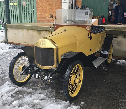 Yellow two-seater with wire wheels and a brass radiator parked on snow by a green shed. Opens a larger view