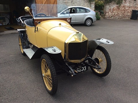 Pale yellow two-seater with a brass radiator and gold wire wheels parked on tarmac by a garage. Opens a larger view