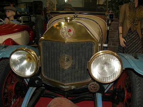 Head-on close view of a brass radiator with a script badge and two large glass headlamps. Opens a larger view