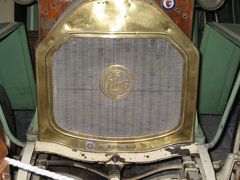 Close view of a brass radiator shell with a script roundel, green wings either side and a rope below. Opens a larger view