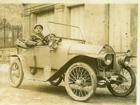 A sepia photograph of a soldier in a steel helmet seated in a small open car with brass lamps before a gateway. Opens a larger view