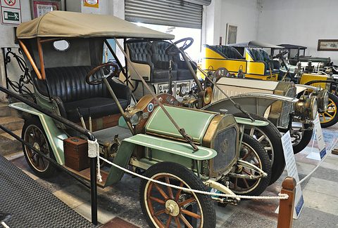 Green two-seater with a tan hood and brass radiator behind a rope barrier, yellow cars behind. Opens a larger view