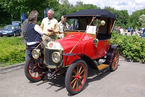 Red two-seater with a black hood and wooden wheels, its owners chatting beside it. Opens a larger view
