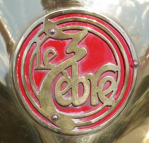 Close-up of a round brass badge with flowing ‘le Zèbre’ lettering on a red background. Opens a larger view