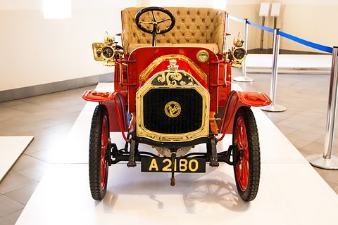 Front of a red two-seater with a gold buttoned seat, brass radiator and red wheels on a white plinth. Opens a larger view