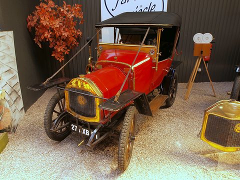 Red single-cylinder two-seater with a brass radiator and black hood on gravel before a dark wall. Opens a larger view