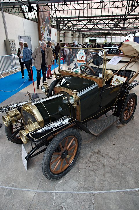 Dark green two-seater with brass lamps under a canopy roof at an indoor show, visitors behind. Opens a larger view